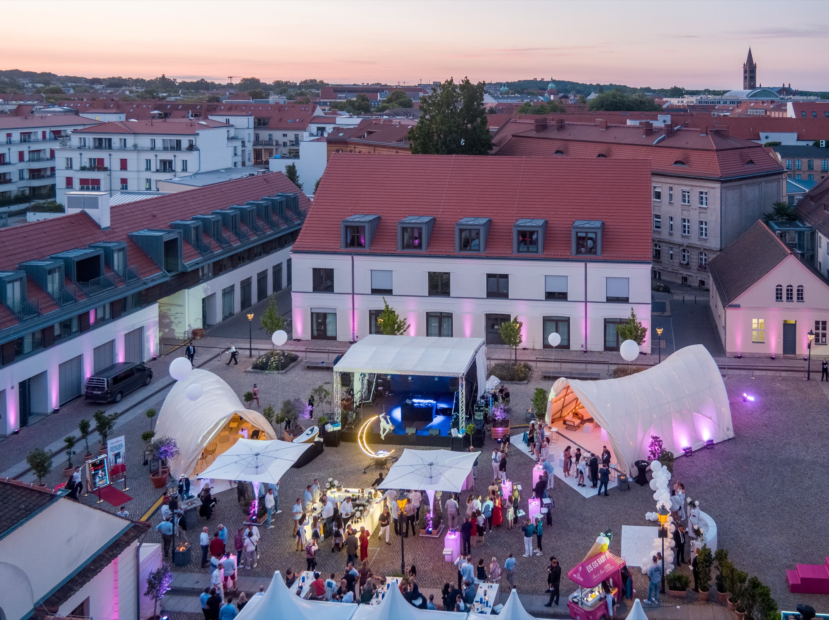 Der Innenhof von oben zur blauen Stunde, Bühne mit Lichtanlage, zwei geschwungene Zelte und Sonnenschirme, dicht besetzte Fläche und der pinke Eiswagen, dahinter die Dächer von Potsdam