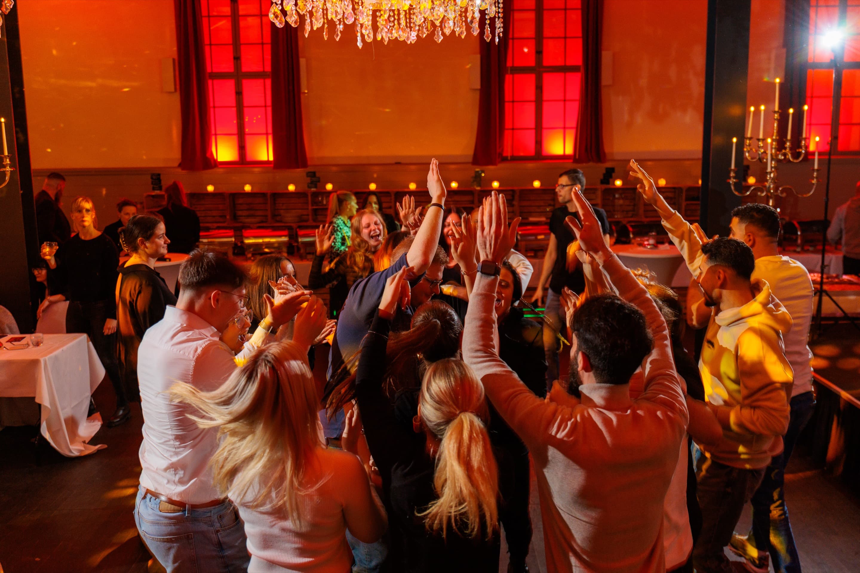 Volle Tanzfläche im Saal, Gäste mit erhobenen Armen unter dem Kronleuchter, warmes rotes Licht