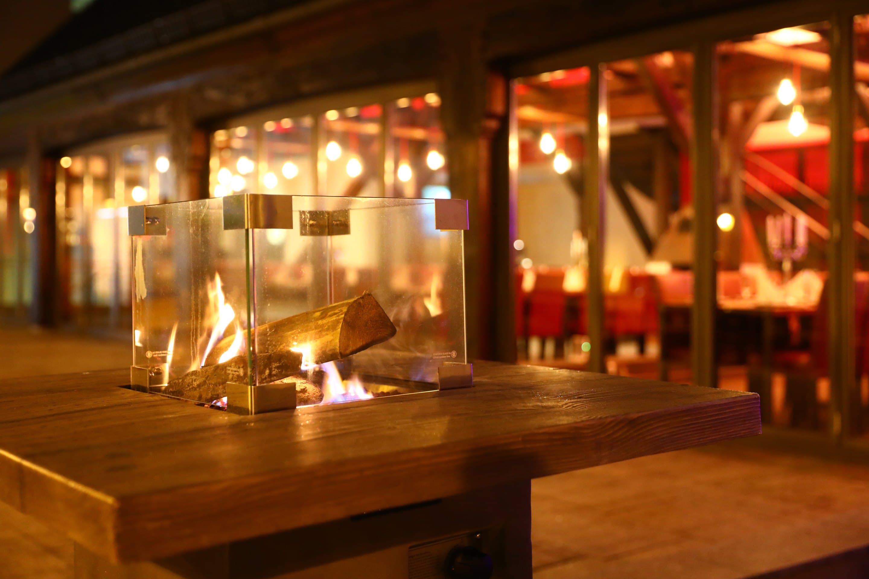 Offenes Feuer in einem Glaswindschutz auf einem Holztisch im Freien, dahinter der beleuchtete Innenraum mit Lichterketten