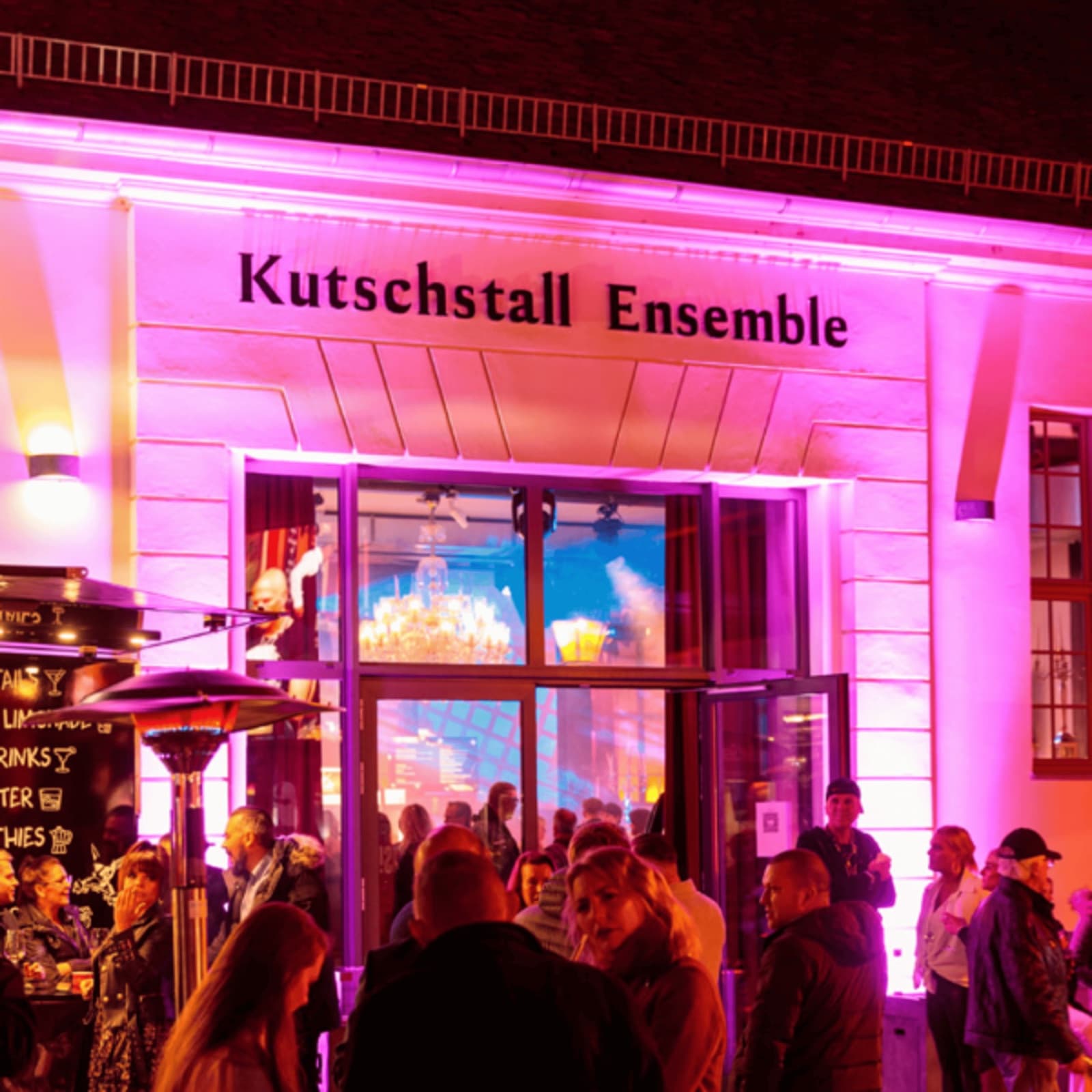 Der Schriftzug Kutschstall Ensemble an der pink angestrahlten Fassade, davor Gäste am Eingang bei Nacht