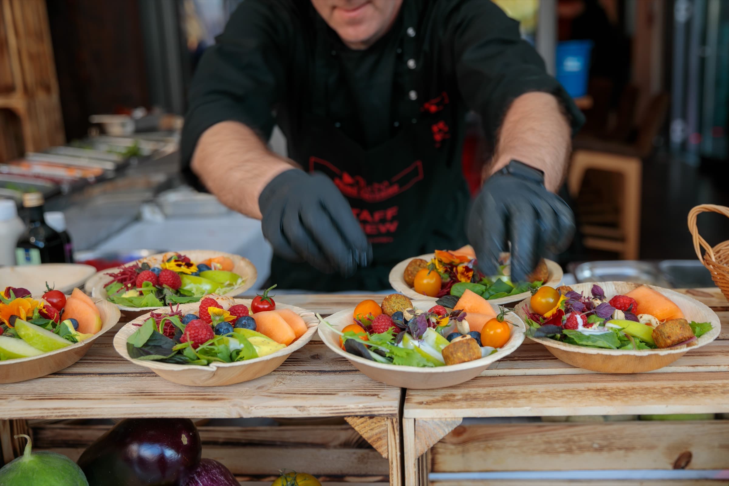 Ein Koch mit schwarzen Handschuhen richtet eine Reihe von Schalen an, darin Blattsalate, Beeren, Melone, Kirschtomaten und essbare Blüten