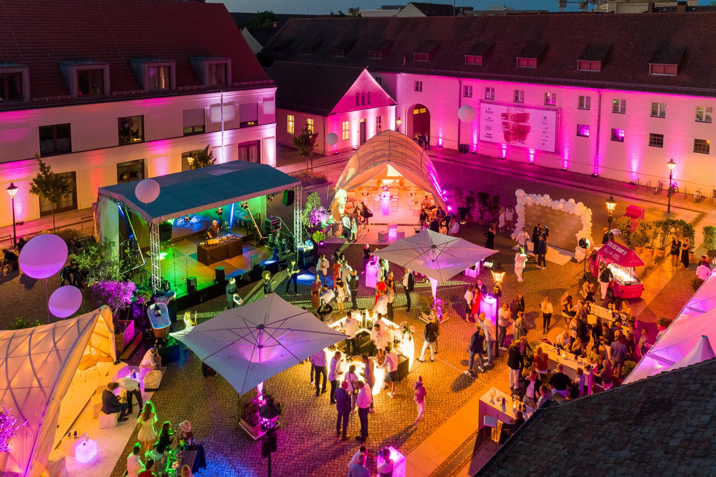 Der Innenhof am Abend bei einem Sommerfest, die historischen Gebäude in Pink angestrahlt, Bühne, Zelte und Foodtruck zwischen Gästen an Stehtischen