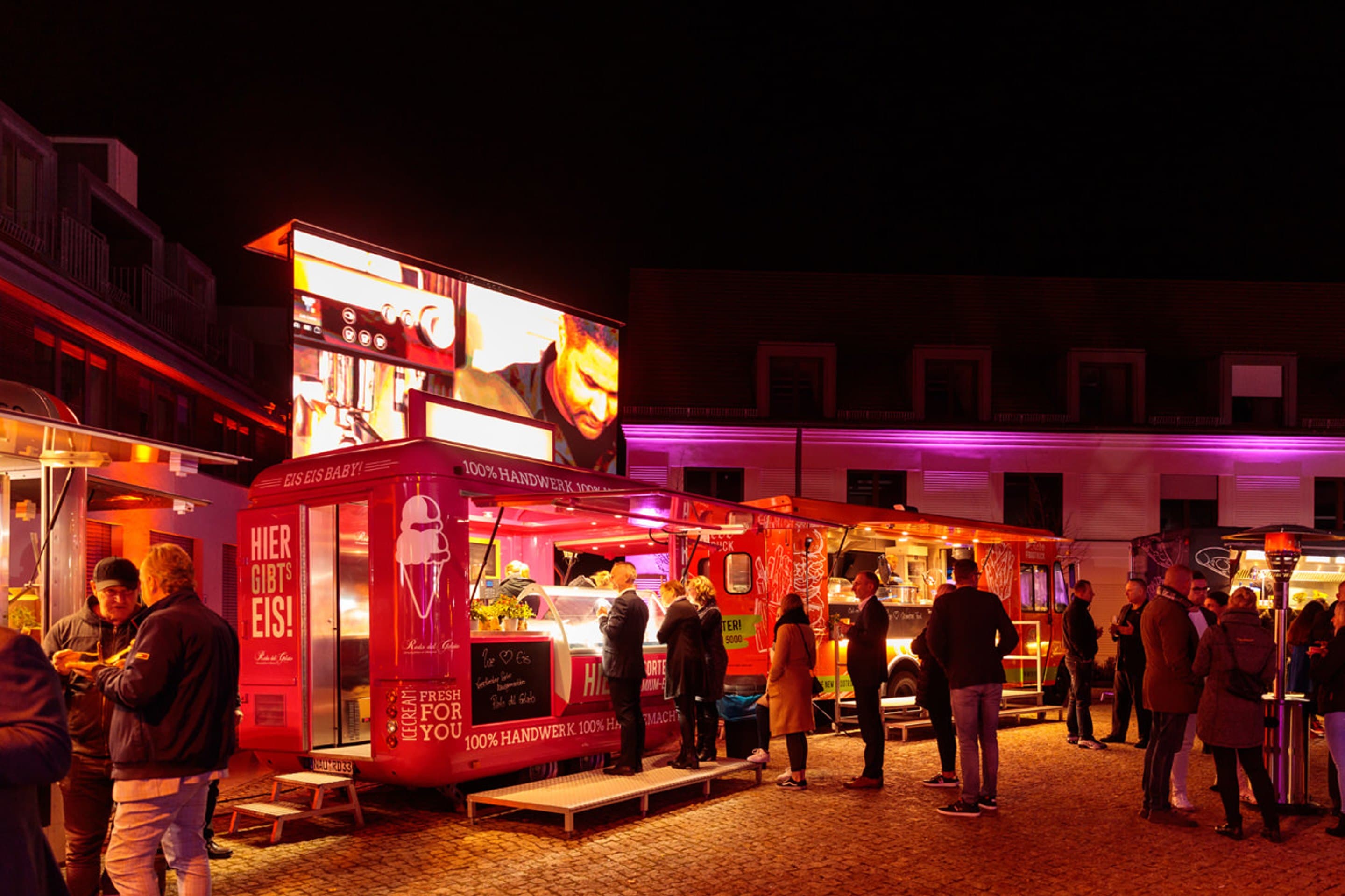 Foodtrucks abends im Innenhof, vorn der rote Eiswagen Eis Eis Baby, Gäste stehen an den Ausgabetheken