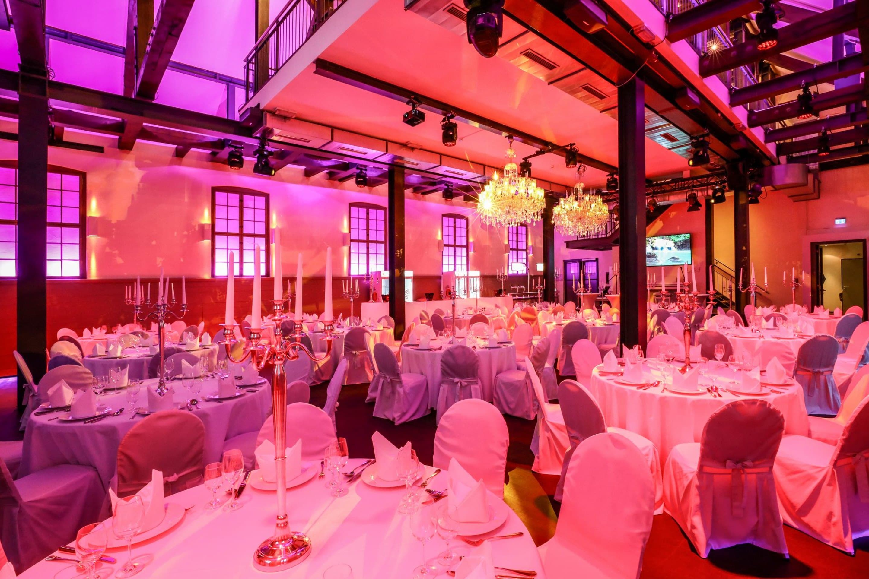 Der Saal für ein Bankett eingedeckt, runde Tische mit weißen Hussen und silbernen Kerzenleuchtern, zwei Kronleuchter und Empore, Wände in Pink angestrahlt